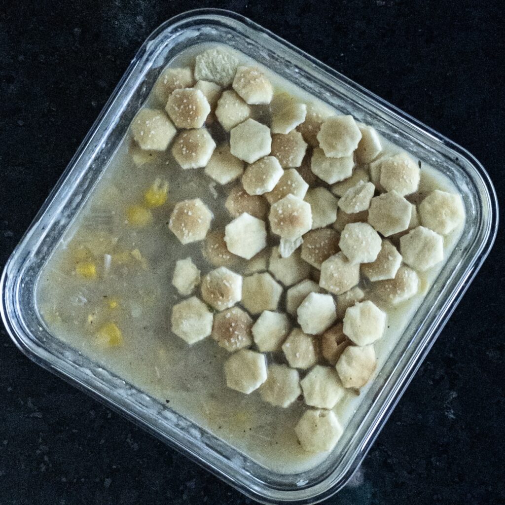 Picture of square container with chowder in it and topped with oyster crackers.