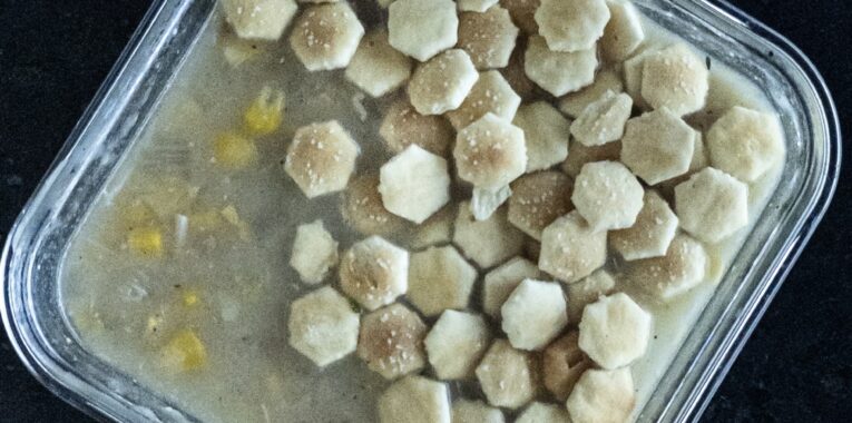 Picture of square container with chowder in it and topped with oyster crackers.