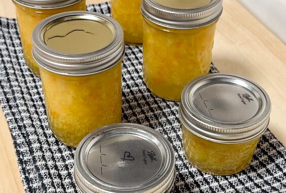 Image of 6 mason jars sitting on a black and white kitchen towel with yellow filling. The filling is pineapple habanero jam. One jar at the front has a heart on it, marked in sharpie.