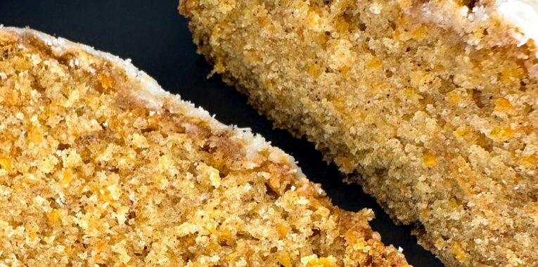 Picture is a loaf of carrot cake with a slice cut off. They are pictured on a black plate.