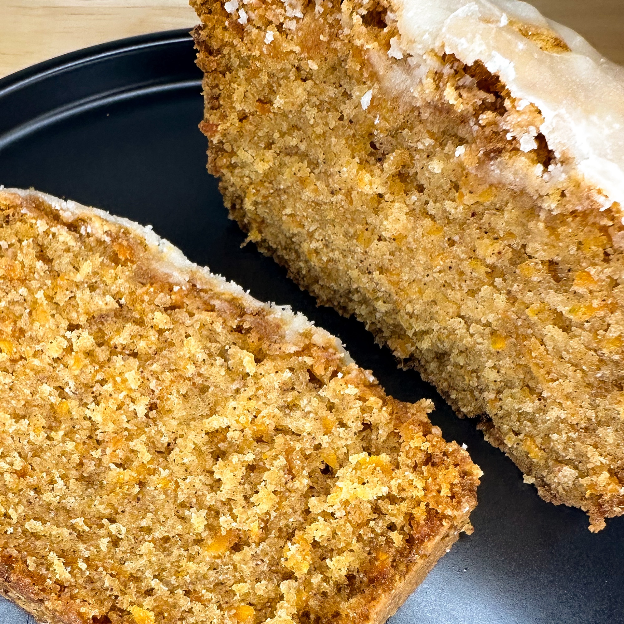 Picture is a loaf of carrot cake with a slice cut off. They are pictured on a black plate.