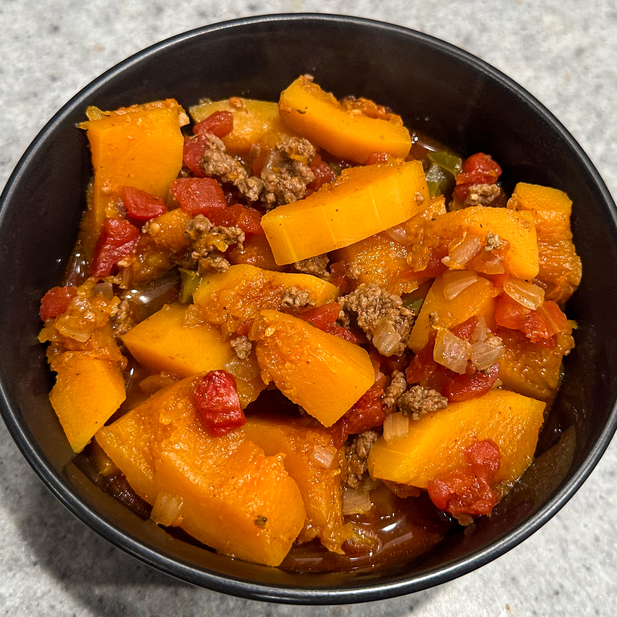 Image is a black bowl with chili in it. The chili has diced tomatoes, ground meat, and butternut squash pieces in it.