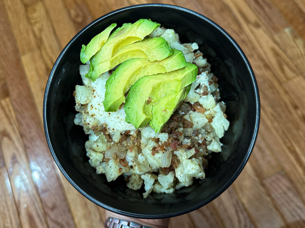 Image of a bowl with food. The food appears to be cauliflower, bacon, and eggs, topped with avocado slices.