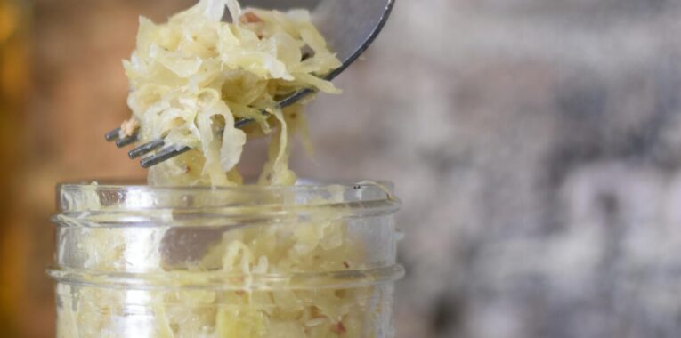 Image of a jar containing fermented cabbage or sauerkraut. An un-pictured individual is holding a fork above the jar with some sauerkraut on it.